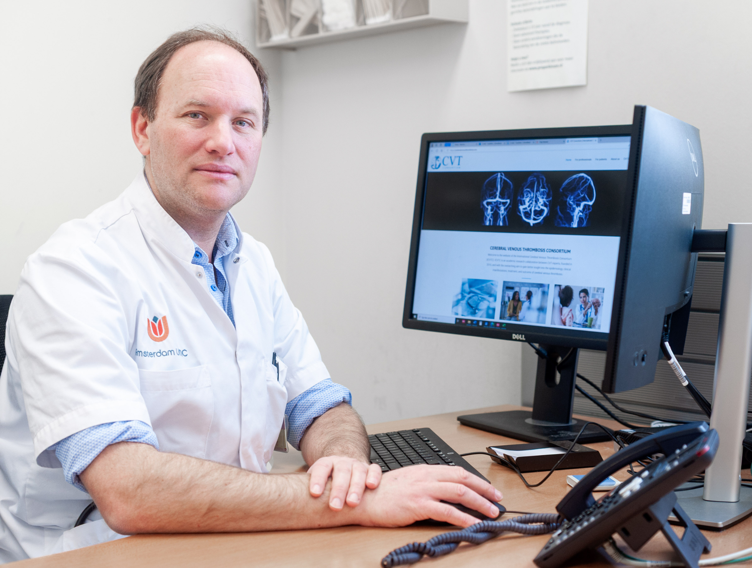 Prof. dr. Jonathan Coutinho Arts in witte jas zit aan een bureau en kijkt in de camera, met een computerscherm op de achtergrond waarop medische beelden en informatie zichtbaar zijn.